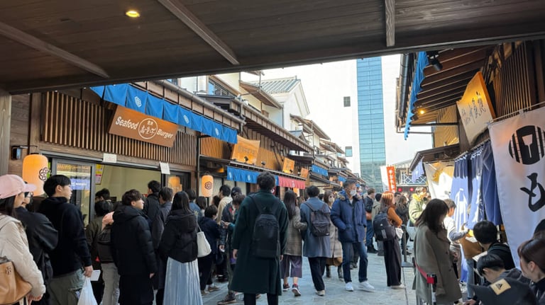 A crowded street at Toyosu Senkyaku Banrai with visitors lining up at storefronts, including a "Seafood Burger" shop. Traditional wooden architecture with blue awnings and hanging lanterns.