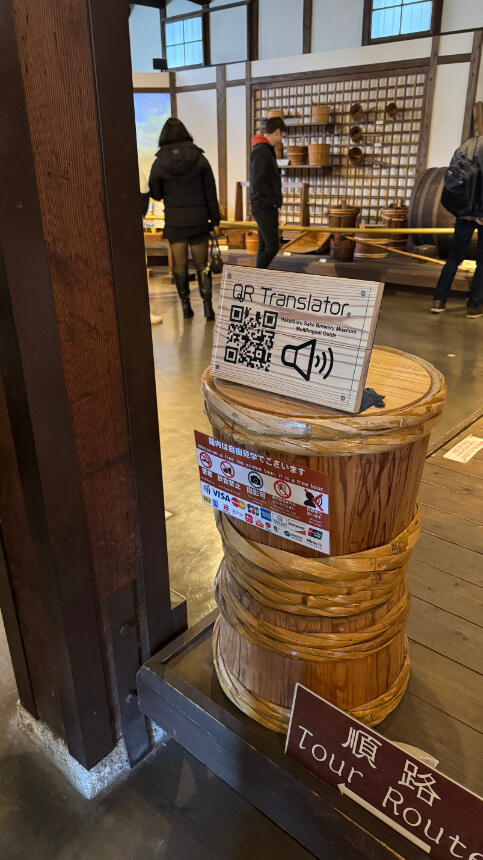 QR Translator sign atop wooden barrels at Hakutsuru Sake Brewery Museum, offering audio descriptions.