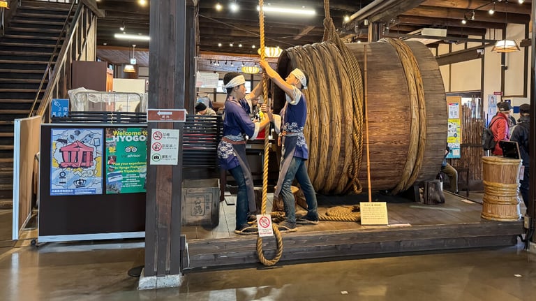 Mannequins in traditional attire demonstrating barrel lifting with thick ropes, with a massive wooden barrel behind them.