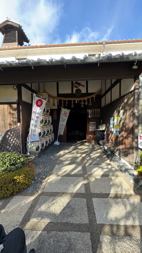 Entrance to Hakutsuru Sake Brewery Museum with flat stone pathway, kura-style facade, banners, and decorative sake barrels. No tactile pavement.