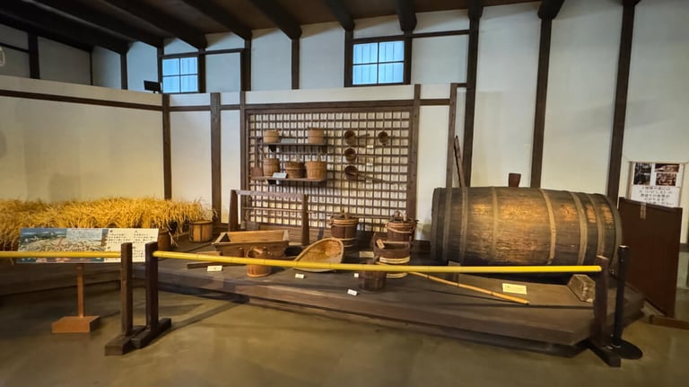 Display of traditional sake-making tools, including barrels and baskets, with a low yellow railing allowing easy viewing from a wheelchair.