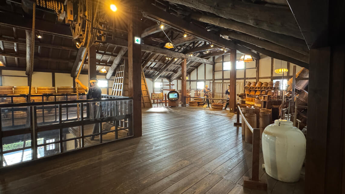 Wooden second floor at Hakutsuru Sake Brewery Museum with displays of brewing tools, mannequins, and rope equipment under a rustic roof.