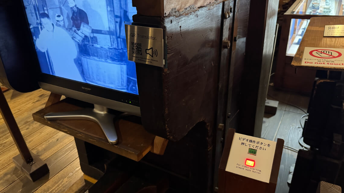 Video display at Hakutsuru Sake Brewery Museum with a button to play content and a QR code for audio translation.