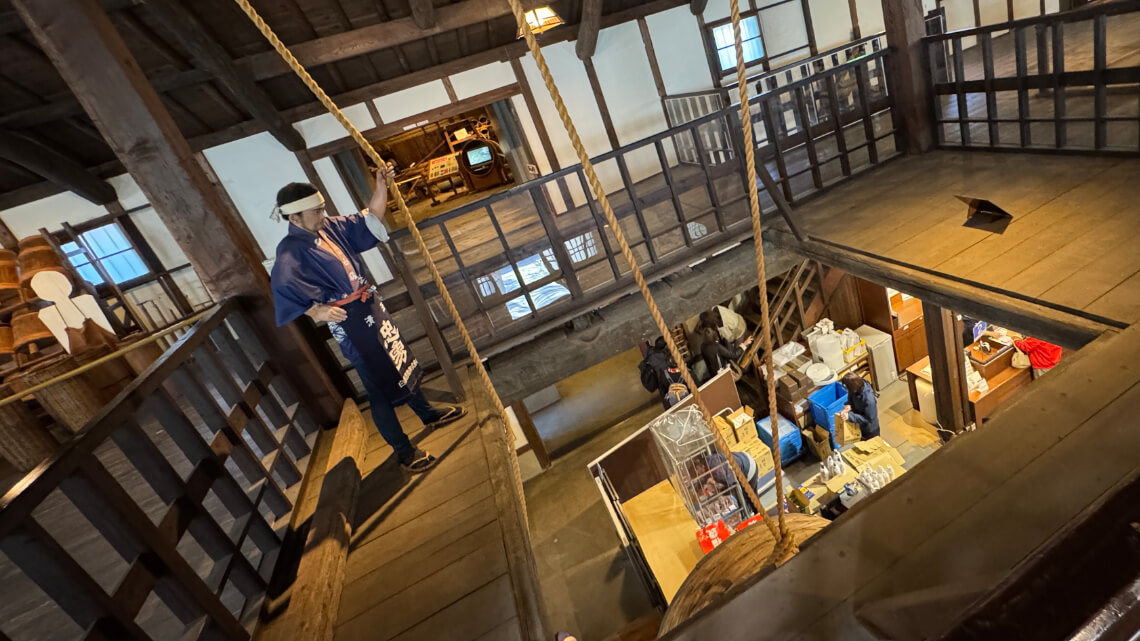 Mannequin on the second floor of Hakutsuru Sake Brewery Museum holding a rope. A cutout in the floor provides a view of the gift shop below.