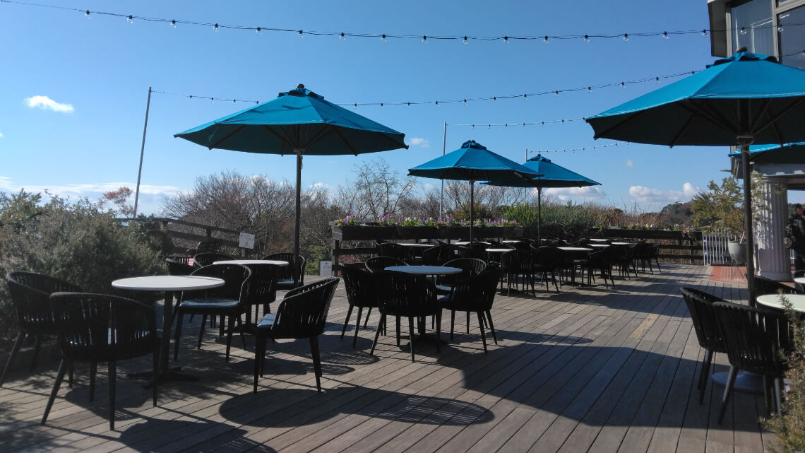 Outdoor rest area at the Glasshouse with tables, chairs, and blue umbrellas on a wooden deck, under string lights and a clear sky.