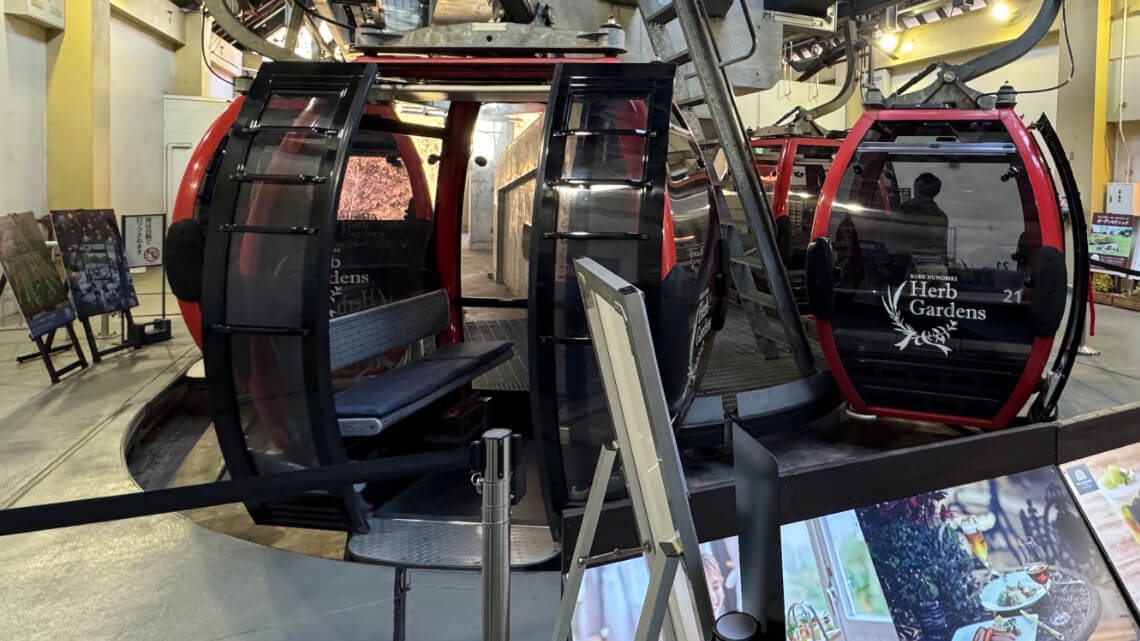 Red cable car gondolas at Kobe Nunobiki Herb Gardens ropeway station, with benches inside and branded glass panels on the sides.