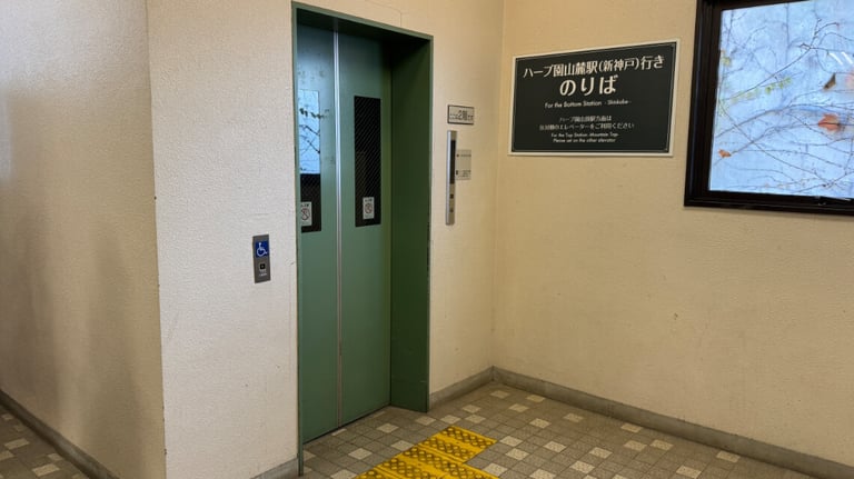 Elevator at the ropeway station with a green door, tactile paving, and a sign indicating access to the bottom station. Elevator at the ropeway station with a green door, tactile paving, and a sign indicating access to the bottom station.
