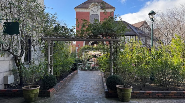 Pathway at Kobe Nunobiki Herb Garden, with greenery, flowerbeds, a wooden archway, and a clock-tower building in view.