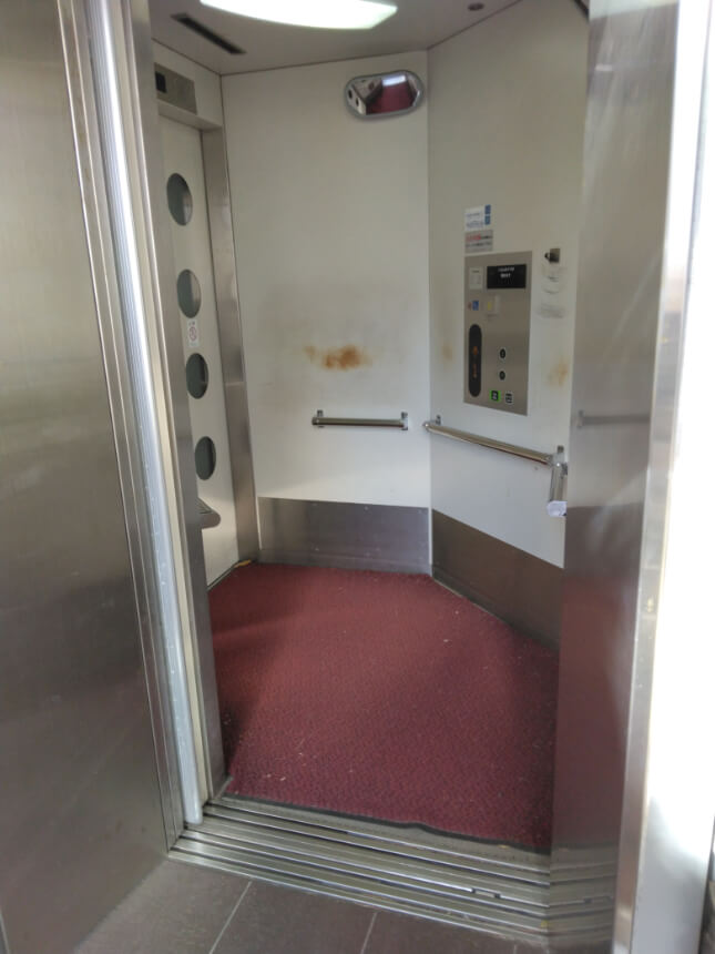Interior of the elevator with low-positioned buttons, a red carpeted floor, handrails, and an exit through the opposite door. Interior of the elevator with low-positioned buttons, a red carpeted floor, handrails, and an exit through the opposite door.