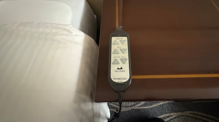 A remote control for an adjustable reclining bed is placed on a bedside table. The buttons are labeled in Japanese - head, feet, and both head and feet.
