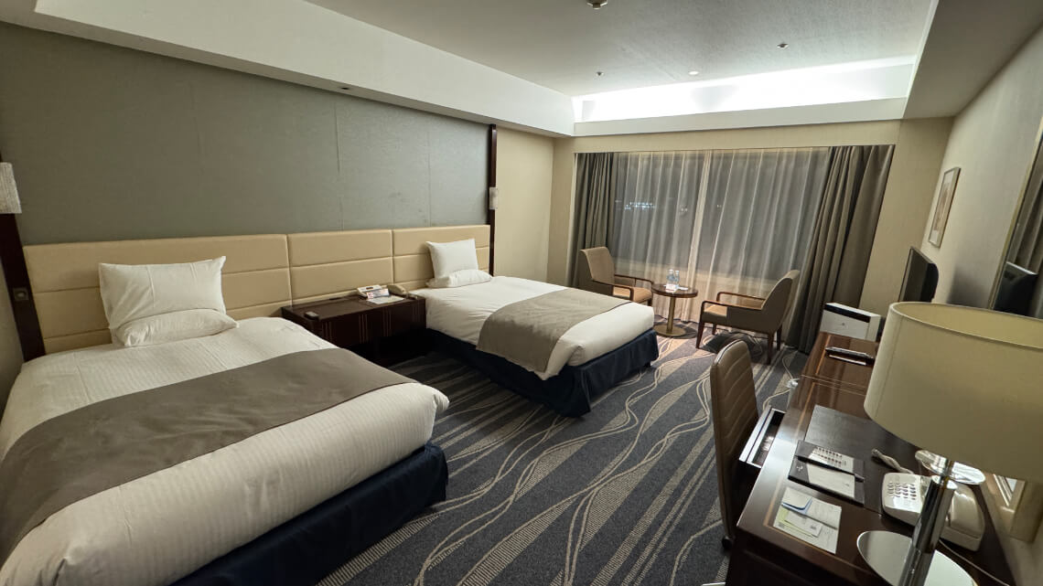 The accessible room features two single beds with neutral bedding, a work desk, seating area by the window, and ample floor space for mobility.
