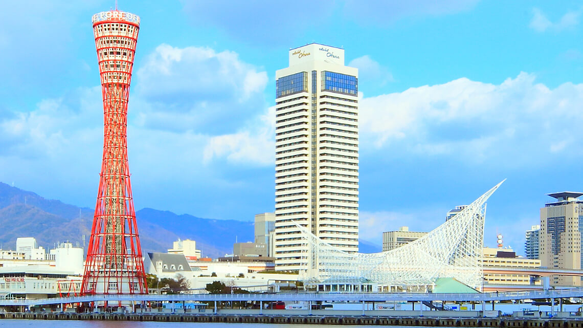 Hotel Okura Kobe stands as a tall, modern building next to the red Kobe Port Tower and the white, angular Kobe Maritime Museum, with a clear sky above.