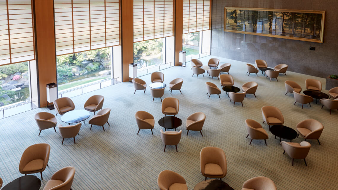 A seating area at Hotel Okura Kobe with circular tables and beige chairs arranged neatly. Large windows overlook a landscaped garden.