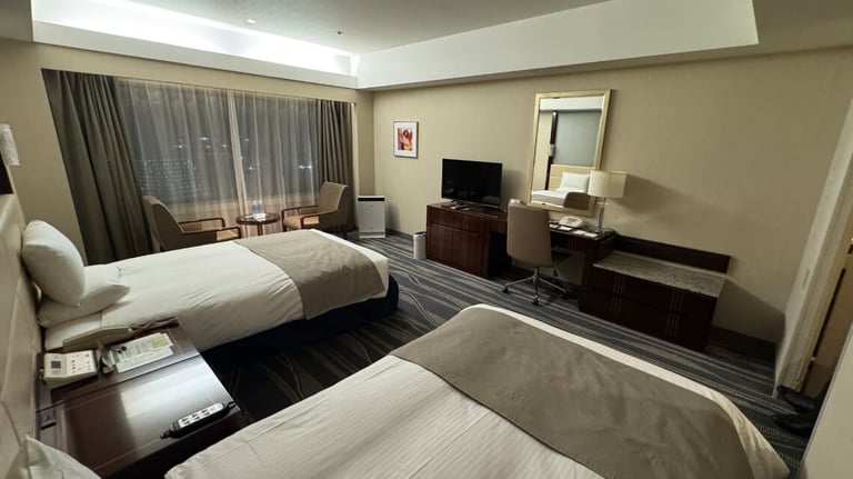 The accessible room includes two single beds, a desk with a chair, a TV on a low cabinet, a seating area by the window, and a wall-mounted mirror.