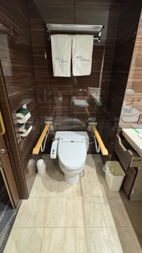 The accessible toilet features a bidet with control buttons, foldable grab bars on both sides, and a towel rack with folded towels above.