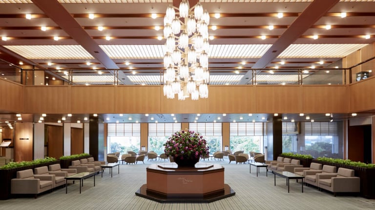 The lobby of Hotel Okura Kobe features a spacious layout with soft seating, a central floral arrangement, warm wood accents, and a large chandelier.