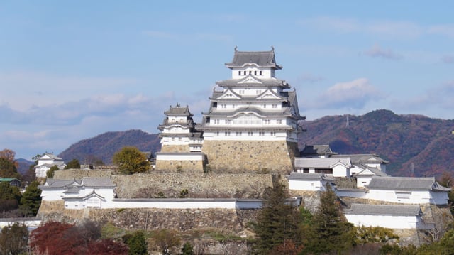Himeji Castle With White Walls And Tiled Roofs, Surrounded By Stone Walls And Trees, Set Against Mountains From The Roof Of A Building Nearby.