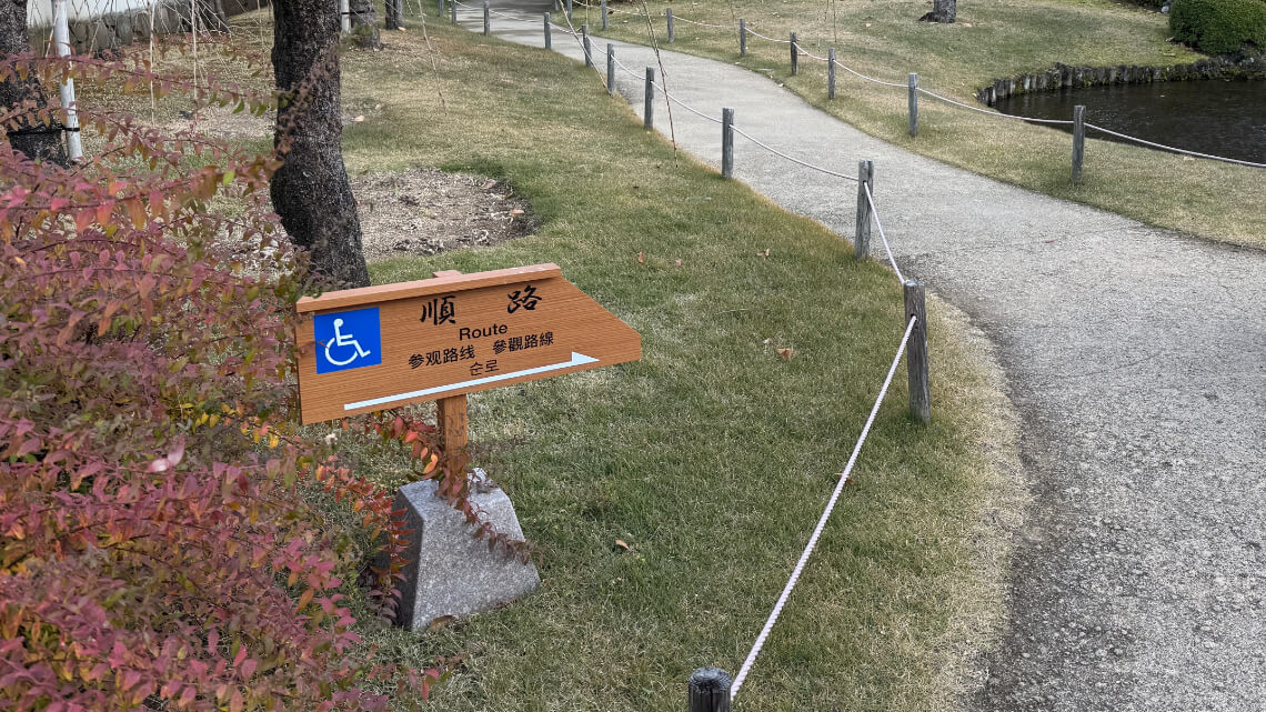 A wooden sign with a wheelchair accessibility symbol marks a smooth pathway bordered by ropes, leading through grass and near a pond.