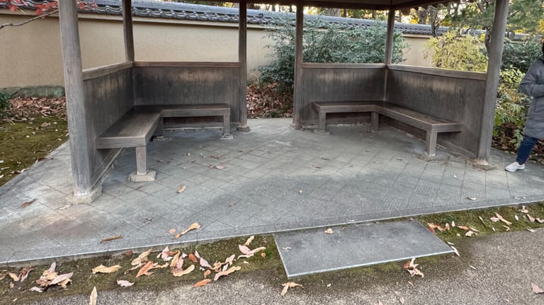 A covered rest area with wooden benches on tiled flooring, with a small step leading from the main garden pathway.