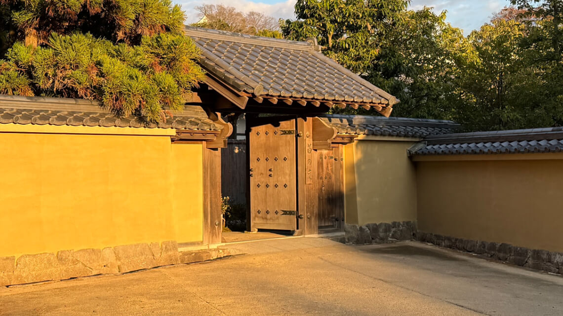 A traditional wooden gate with tiled roofs set in yellow plaster walls. There is a small bump in the pathway leading through the gate.