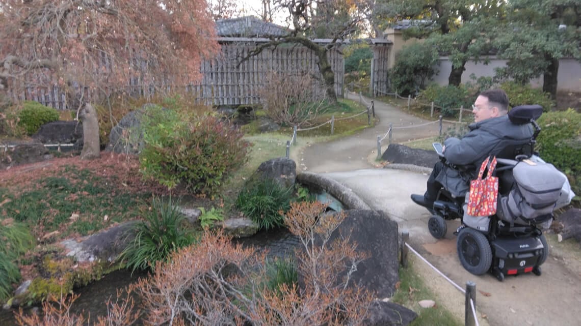 A person in a motorized wheelchair is exploring a peaceful Japanese garden with a small bridge, trees, and a gravel pathway.