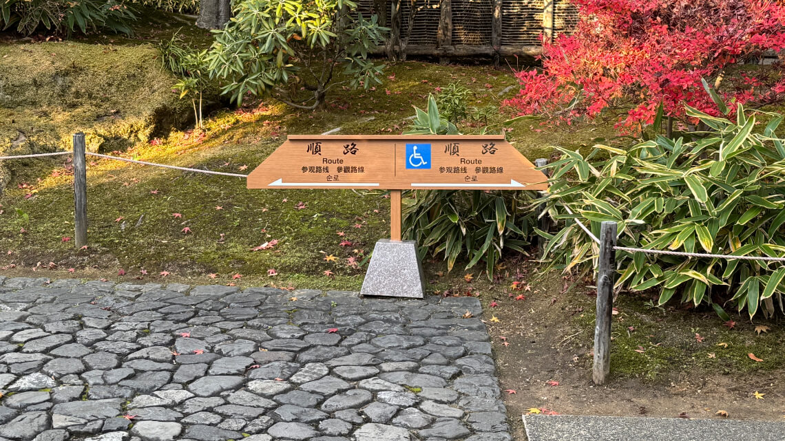 A wooden sign marking routes, including one with a wheelchair accessibility symbol, set on a cobblestone path.