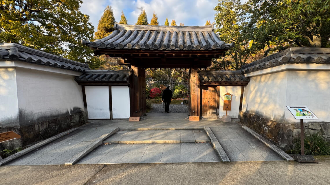 The start of the garden, a traditional wooden gate with tiled roofs, with ramps leading into a garden path surrounded by trees.