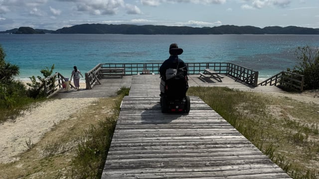 A person in a wheelchair moves along a wooden boardwalk overlooking a scenic ocean view, while another person walks toward the beach.