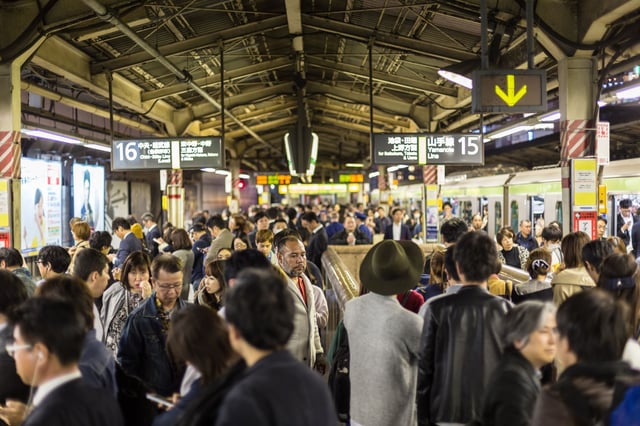 A crowded train platform in Japan with many people waiting, walking, or standing near platforms 15 and 16. Overhead signs in Japanese and English indicate directions for the Yamanote Line and Chūō Line. A train is stopped at the platform on the right, with passengers entering and exiting.