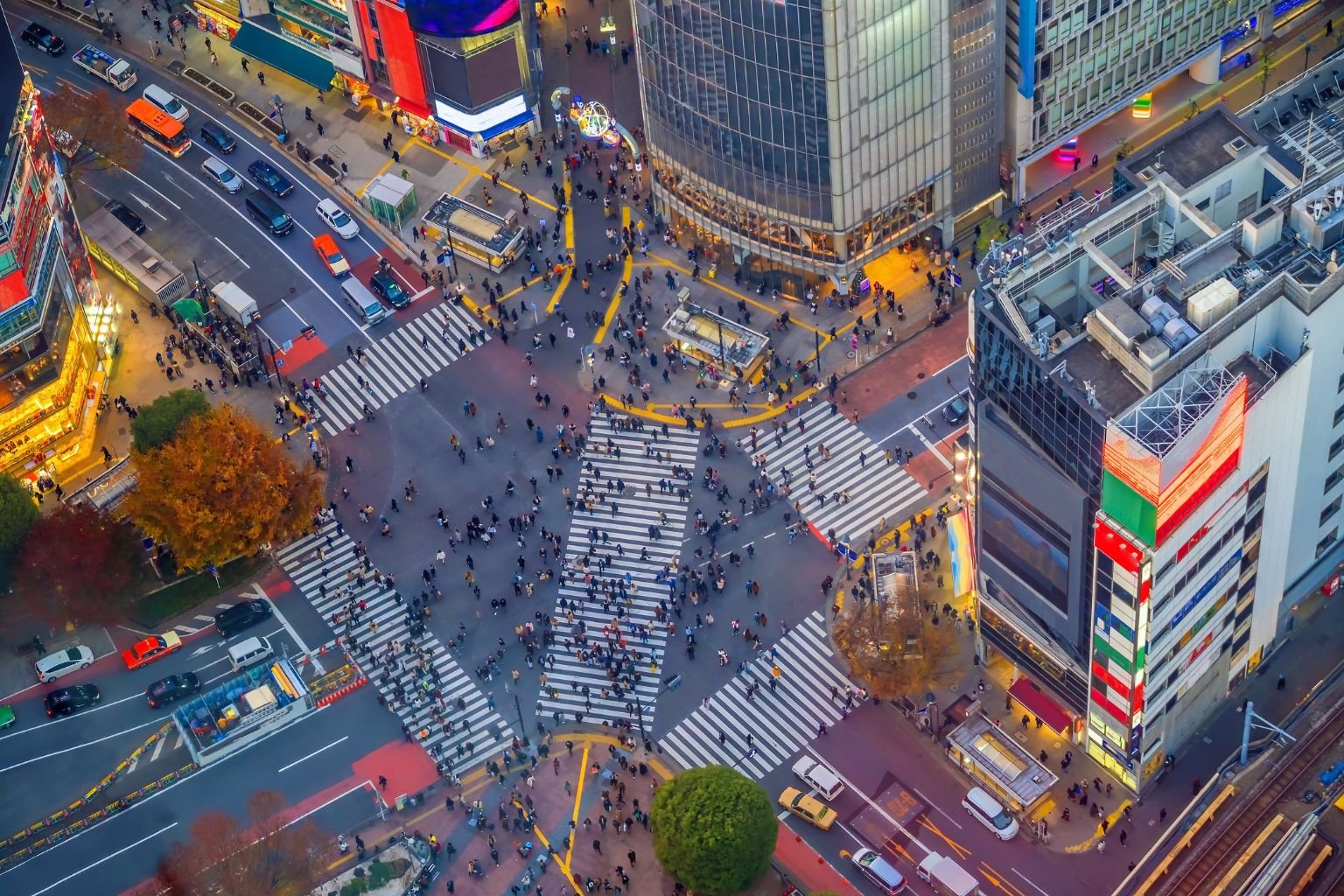 Aerial view of the famous Shibuya Crossing in Tokyo, Japan, with hundreds of people crossing in multiple directions. The intersection is surrounded by tall buildings, colorful billboards, and bright city lights, with cars and buses waiting at the edges.