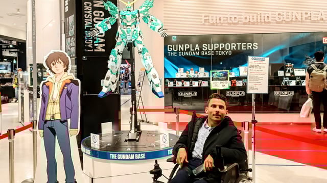JeanFred, a man in a wheelchair, poses in front of a Unicorn Gundam display at The Gundam Base Tokyo, near a cardboard cutout of an anime character.