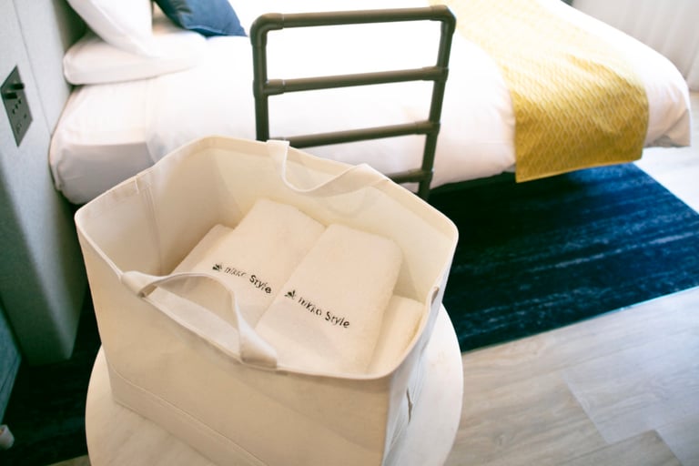 Canvas bag with Nikko Style-branded towels placed on a small table next to an accessible bed with assist rail.
