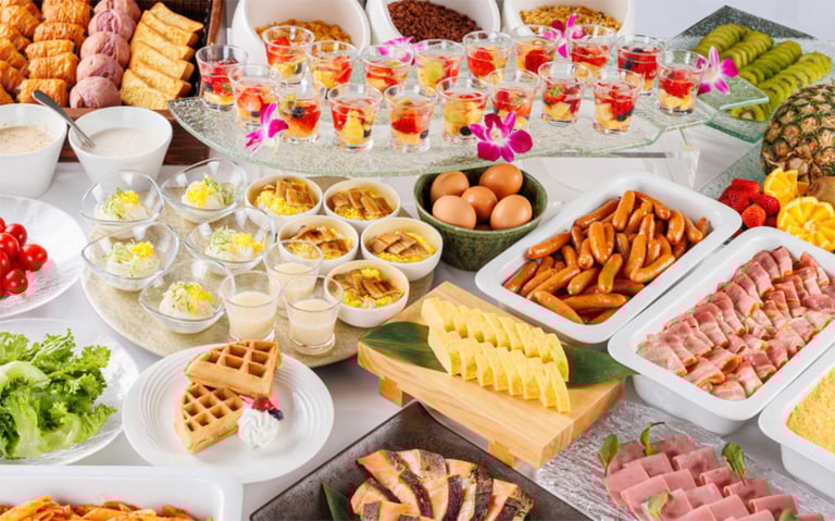Colorful breakfast buffet with eggs, sausages, waffles, fruits, salads, and desserts arranged neatly on a table.