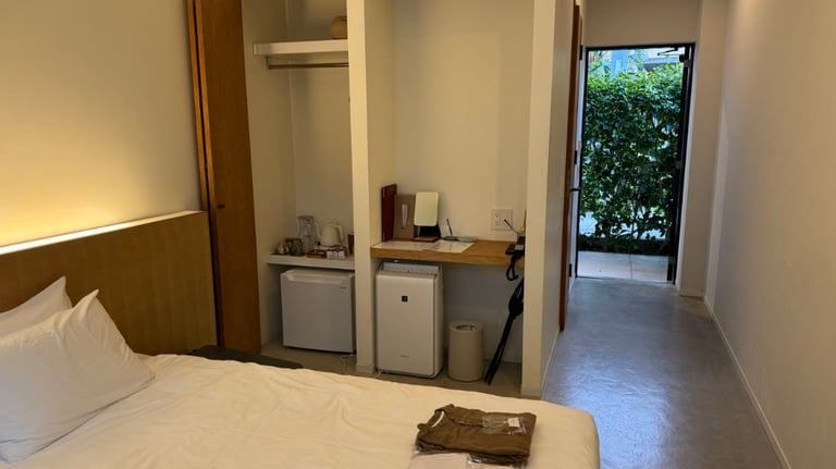 View from bed showing a mini fridge, kettle, desk, and open entryway with a small step leading outside.