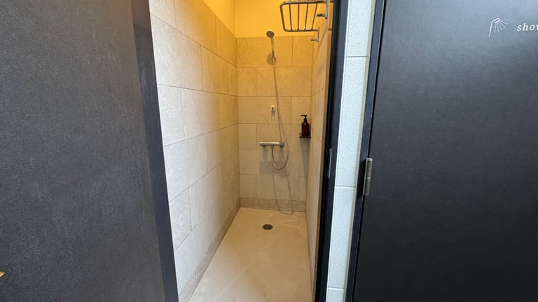 Open black door reveals a narrow tiled shower room with a handheld showerhead, shelf, and wall-mounted soap dispenser.