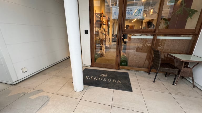 Close-up of Kanusuba entrance showing a glass door, welcome mat, white column, and a small outdoor table with two chairs.