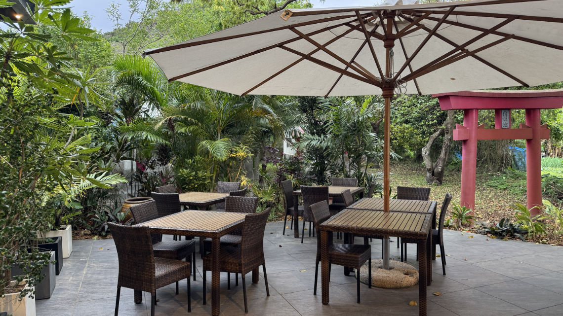 Wicker patio tables under a large umbrella surrounded by greenery, with a red torii gate visible in the background.