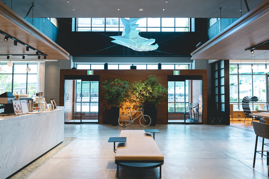 Hotel lobby with high ceilings, a central bench, indoor plants, a white bicycle, and a café counter on the left.