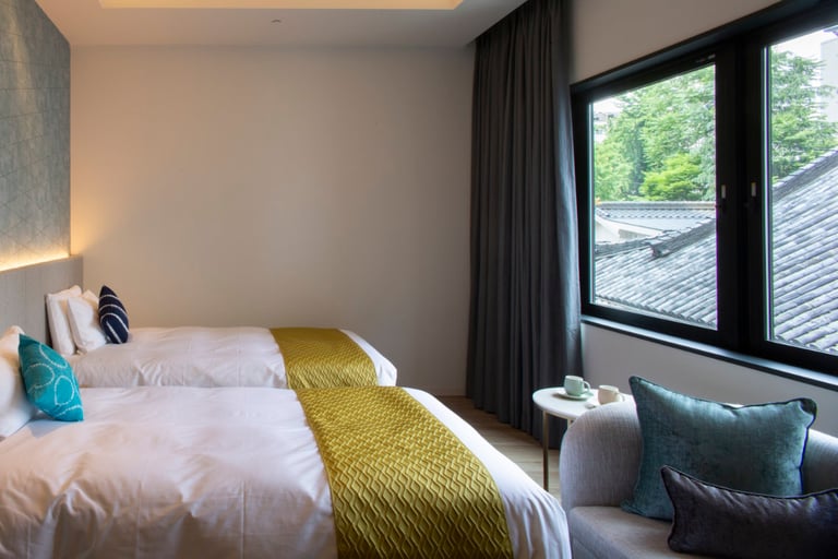 Twin beds with green runners, a window-side seat, and a view of traditional tiled rooftops and trees outside.