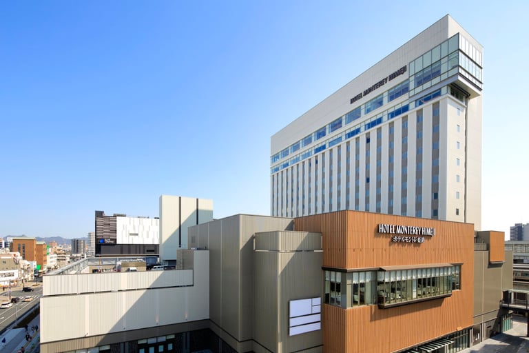 Hotel Monterey Himeji’s modern high-rise exterior under a clear blue sky, with city buildings and roads nearby.