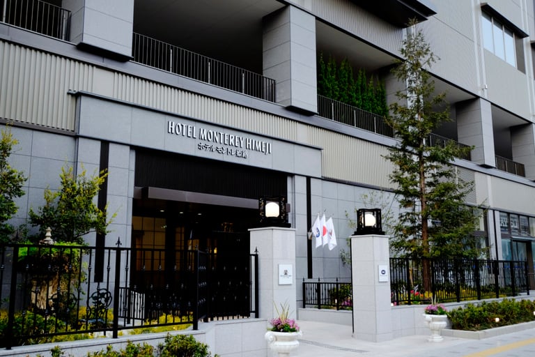 Entrance of Hotel Monterey Himeji with modern stone facade, flags, trees, and black metal fencing.