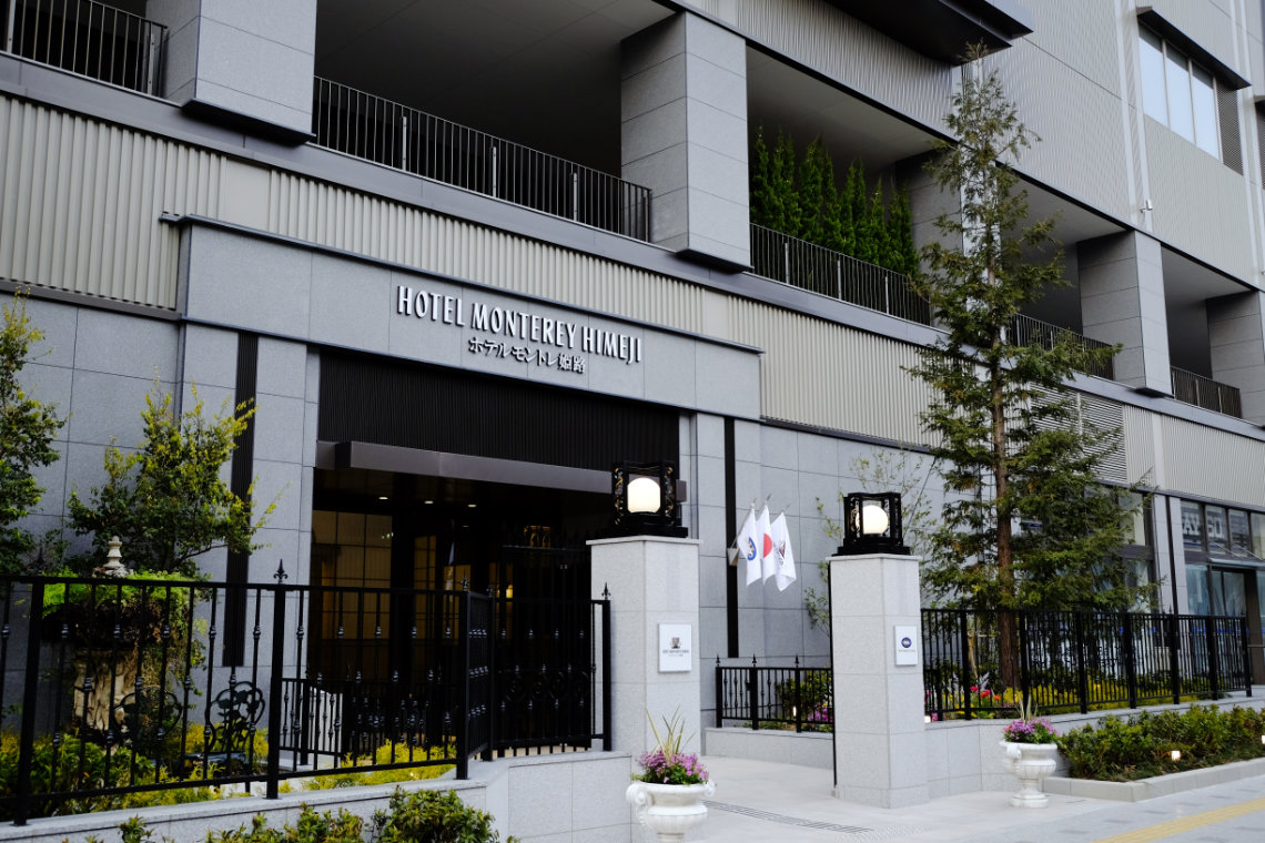 Entrance of Hotel Monterey Himeji with modern stone facade, flags, trees, and black metal fencing.