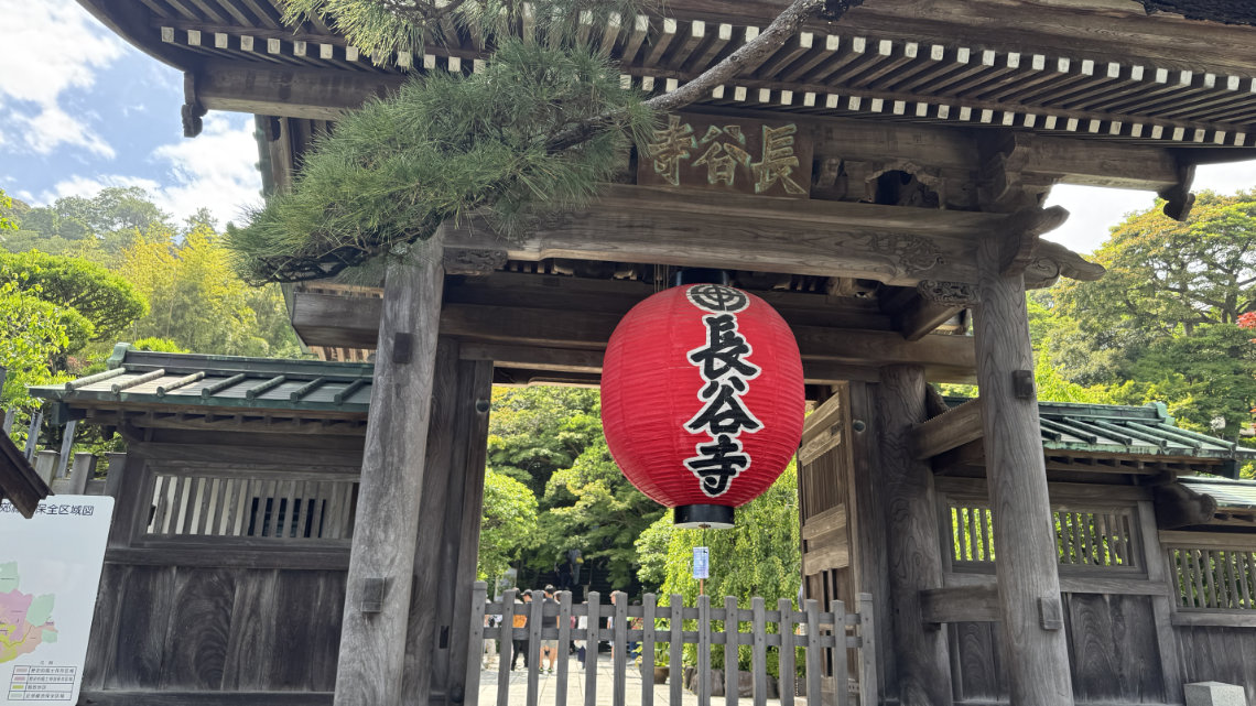 A large red lantern hangs at the wooden gate entrance of Hasedera, surrounded by lush greenery and blue sky.