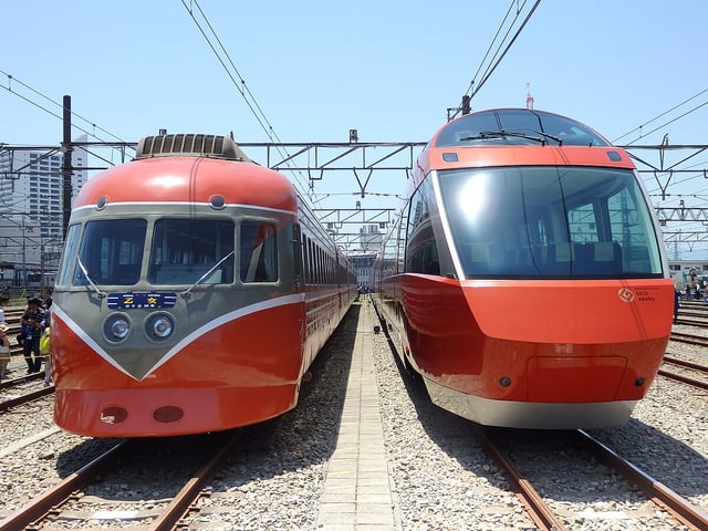 Side-by-side view of vintage and modern Odakyu Romancecar trains on parallel tracks under clear skies.
