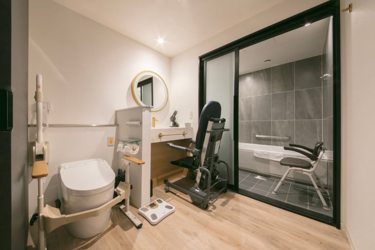 Accessible hotel bathroom with a bidet toilet, assistive rails, shower chair, reclining support chair, and a roll-under sink.