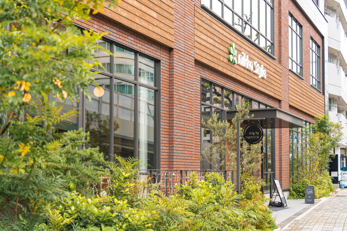Street view of Nikko Style Nagoya with brick exterior, large windows, greenery, and the entrance to Style Kitchen restaurant.