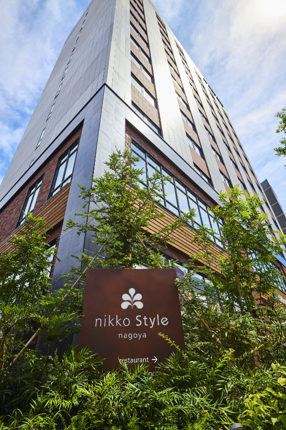 Hotel building with large windows, viewed from below, with a Nikko Style Nagoya sign surrounded by greenery.