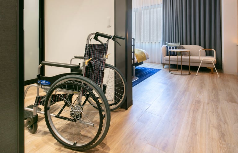 Accessible hotel room entrance with a wheelchair, wide doorway, wooden flooring, and a view of a bed and seating area.
