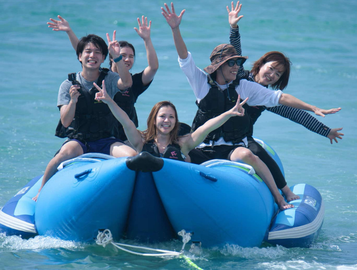 Five people wearing life vests enjoy a banana boat ride, smiling and posing with peace signs on the ocean at Yuimaru Beach Festival.