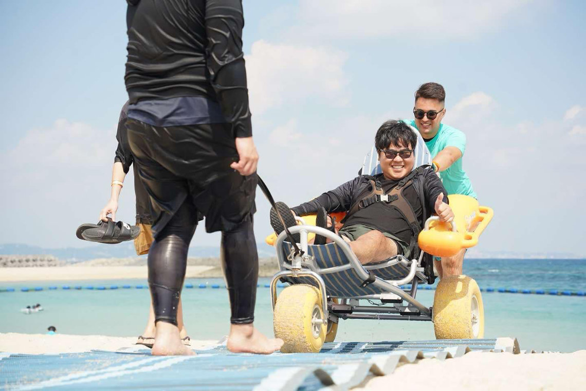 Man in a beach wheelchair is pushed by a friend along a mat path toward the ocean at Yuimaru Beach Festival.
